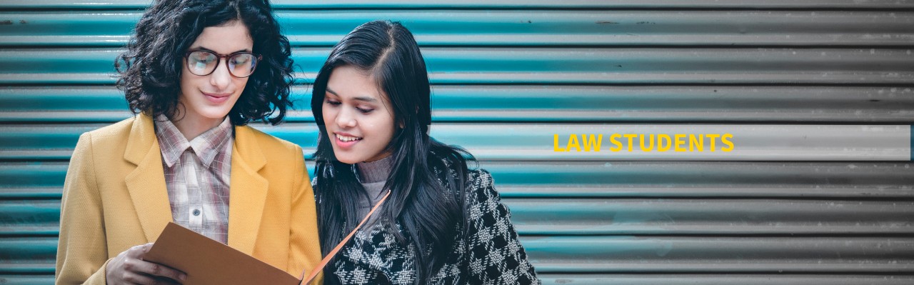 Outdoor image of two happy, Asian/Indian young adult businesswomen, stand against a closed shutter of commercial complex and discuss a project in a folder on the go at day time.